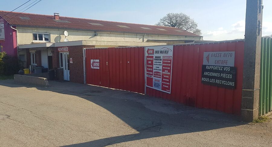 L'entrée de la casse : palissade en tôle rouge, panneaux « Casse Auto Entrée » et « Rapportez vos anciennes pièces, nous les recyclons »